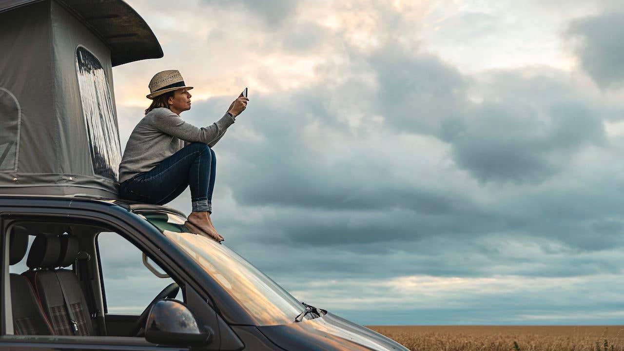 Woman taking a photo whilst sitting on roof of camper at sunset