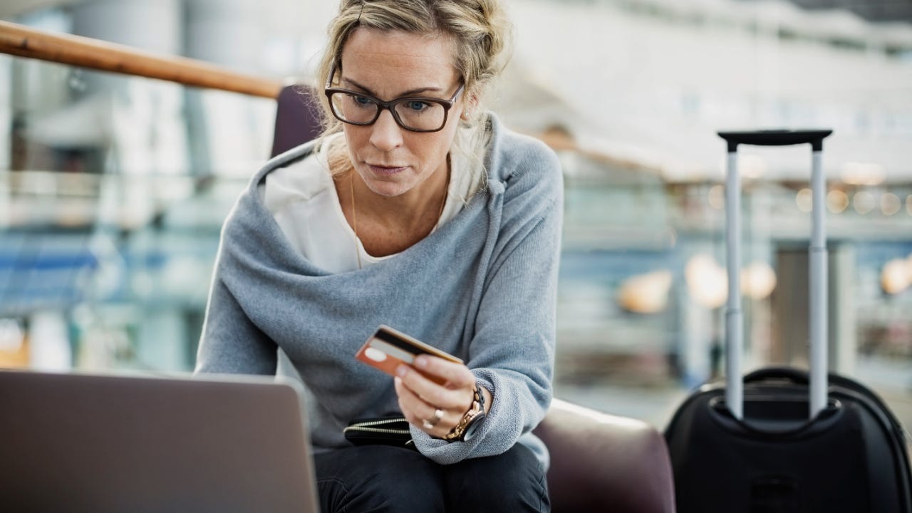 Middle-aged woman with glasses holds credit card while working on laptop with a suitcase nearby