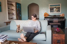 A woman with a laptop sits on a couch with a small dog