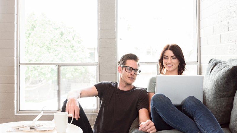 A young couple sits on a couch with their laptop