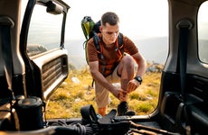 A person in hiking gear with a tee shirt and khaki shorts. They have a backpack on and they are leaning up against the bed of their car (backdoor open) as they check their boot laces.