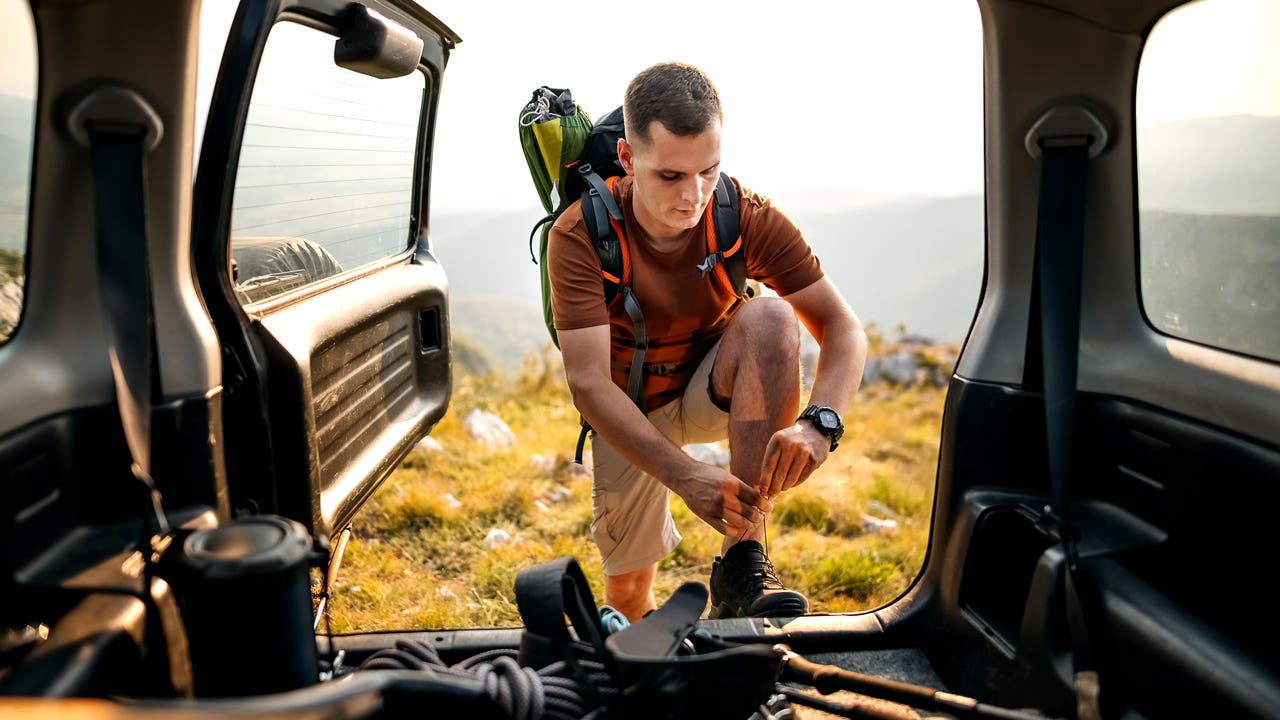 A person in hiking gear with a tee shirt and khaki shorts. They have a backpack on and they are leaning up against the bed of their car (backdoor open) as they check their boot laces.