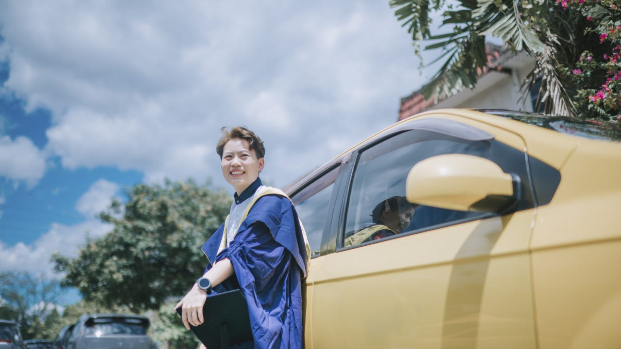 asian chinese young female with graduation gown leaning on her car looking at camera smiling