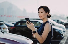 Confident and professional young Asian businesswoman walking to her car in an outdoor carpark in the city, using smartphone and holding a cup of coffee. Business on the go concept
