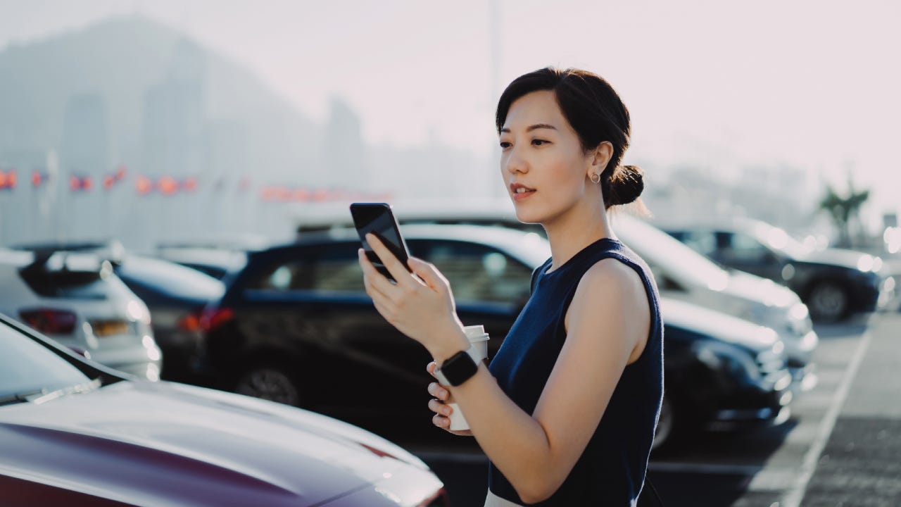 Confident and professional young Asian businesswoman walking to her car in an outdoor carpark in the city, using smartphone and holding a cup of coffee. Business on the go concept