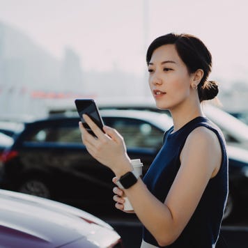 Confident and professional young Asian businesswoman walking to her car in an outdoor carpark in the city, using smartphone and holding a cup of coffee. Business on the go concept