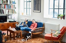 Retired couple sitting on sofa at home, relaxing, woman with laptop smiling, man with head back wearing headphones, lost in music, sitting side by side on blue sofa in retro style living room