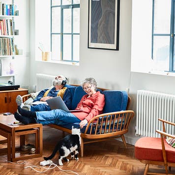 Retired couple sitting on sofa at home, relaxing, woman with laptop smiling, man with head back wearing headphones, lost in music, sitting side by side on blue sofa in retro style living room
