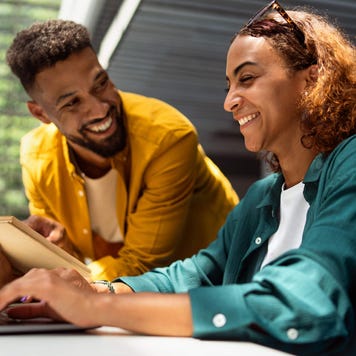 Two university students smile and talk in a library as one holds an open book and the other uses a laptop