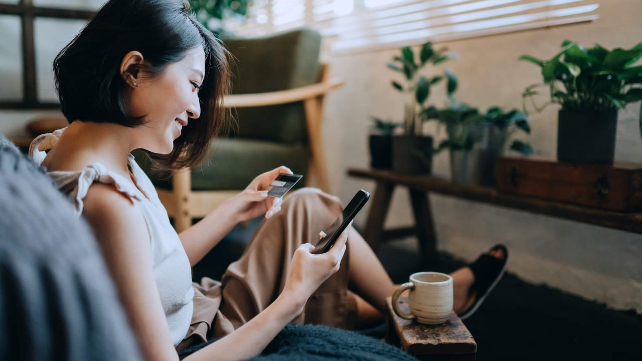 Woman sitting on floor at home shopping online with smartphone while making mobile payment with credit card on hand