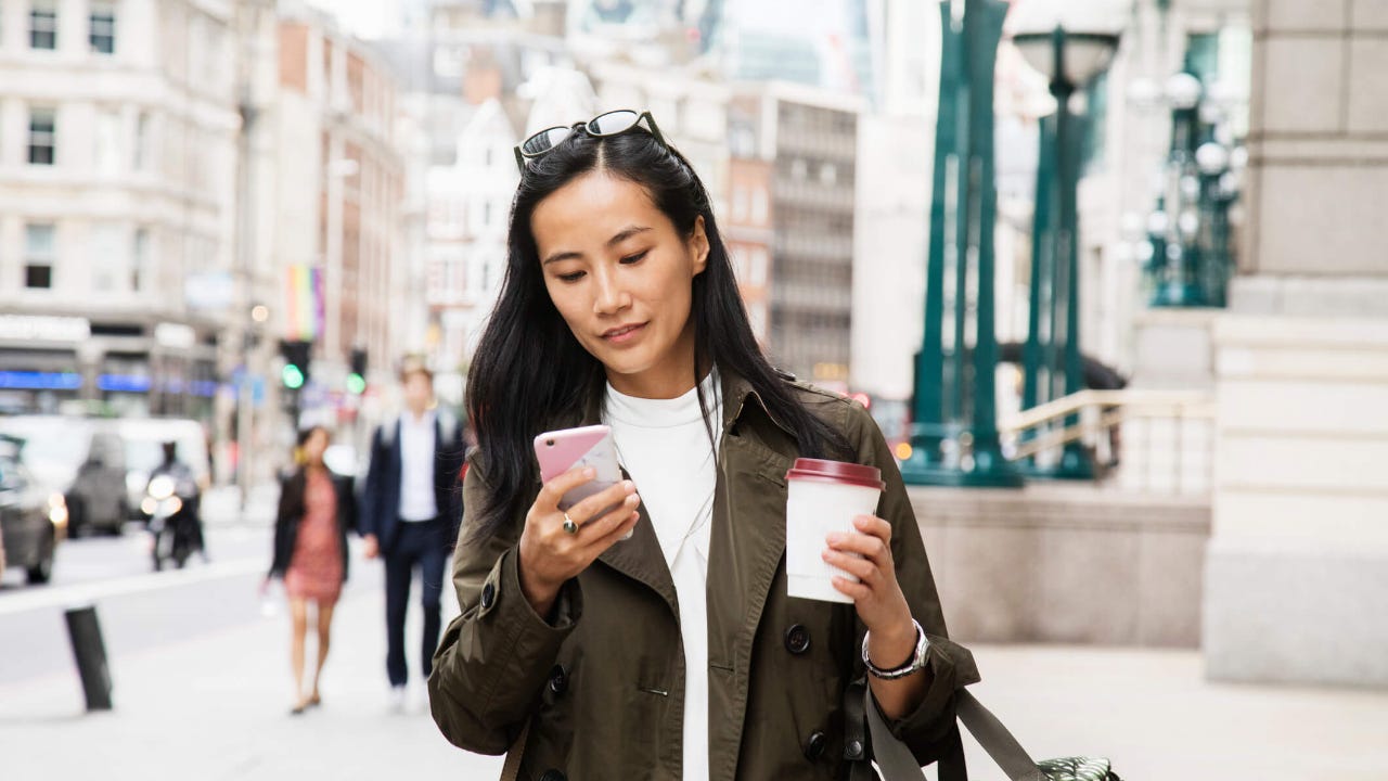 Young woman walks down a city street with coffee while using her smartphone