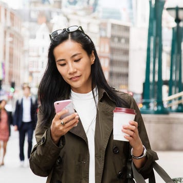 Young woman walks down a city street with coffee while using her smartphone