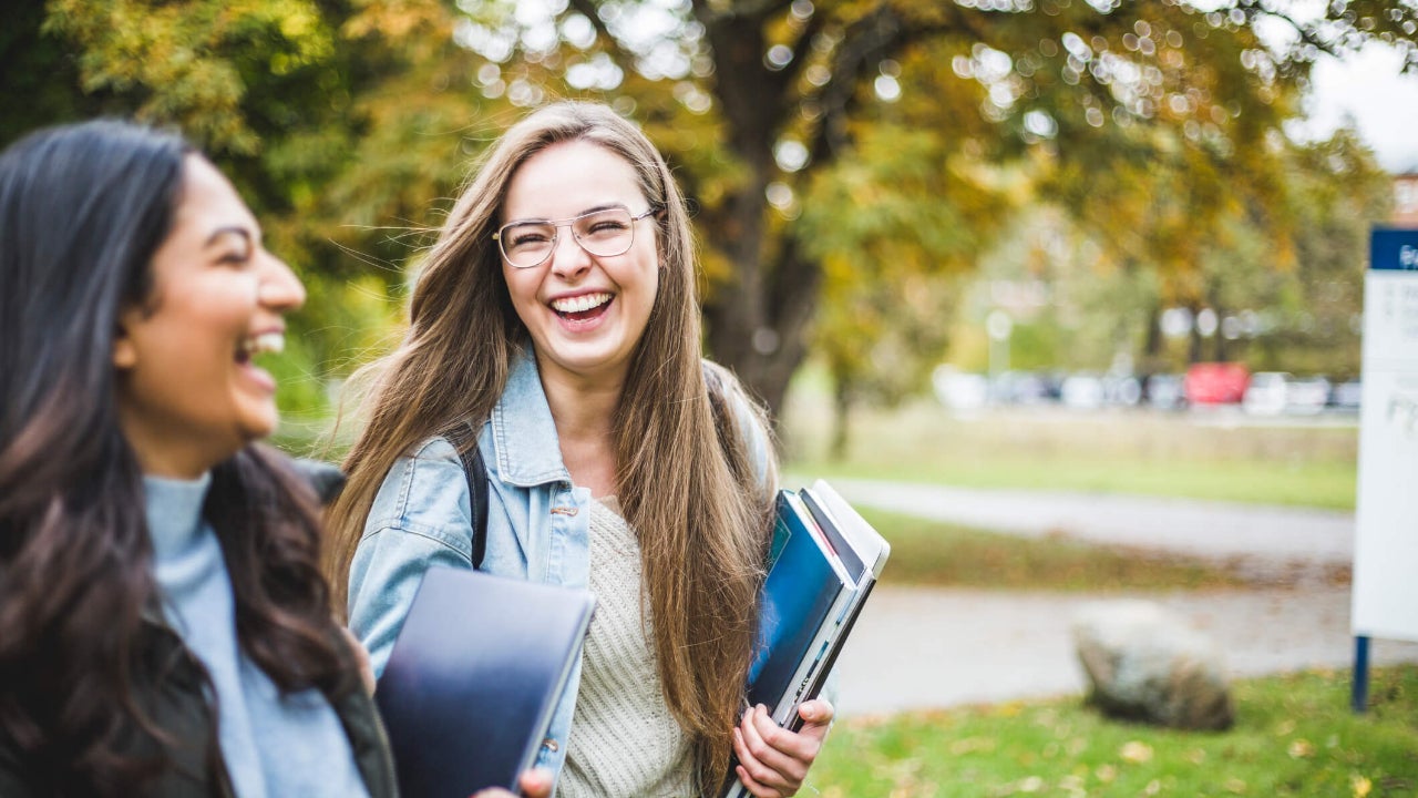 Two happy female students walk and laugh on a university campus with books in hand