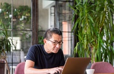 Senior man with glasses uses a laptop at a desk surrounded by house plants
