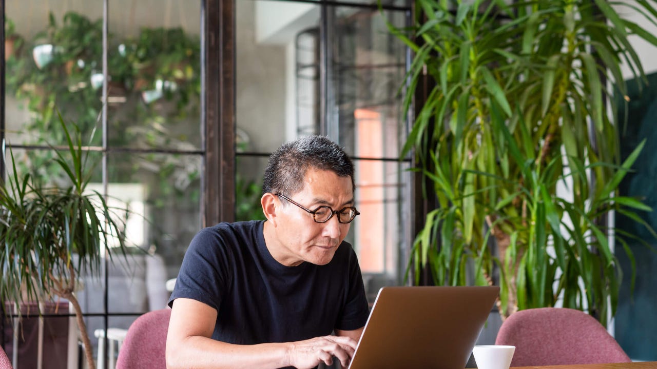 Senior man with glasses uses a laptop at a desk surrounded by house plants