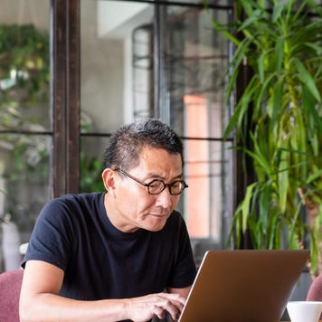 Senior man with glasses uses a laptop at a desk surrounded by house plants