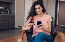 Young woman smiles at her smartphone with credit card in hand in kitchen