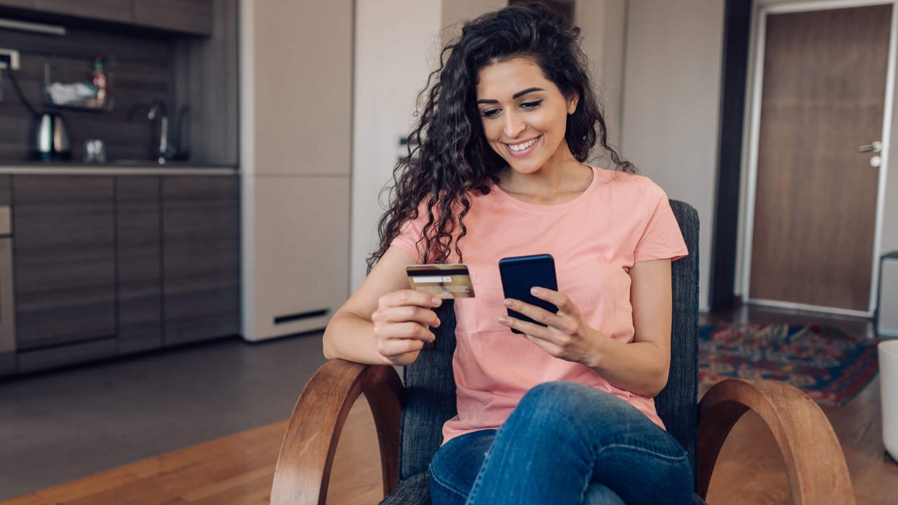 Young woman smiles at her smartphone with credit card in hand in kitchen