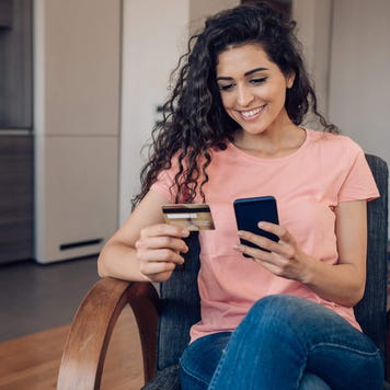 Young woman smiles at her smartphone with credit card in hand in kitchen