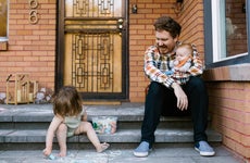 A parent sits on the stoop in front of their home with one baby child in their arms and another, toddler, drawing with chalk on the walkway.