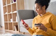 Young woman sits on the floor with her back against a couch as she uses her credit card to make a mobile purchase on her smartphone