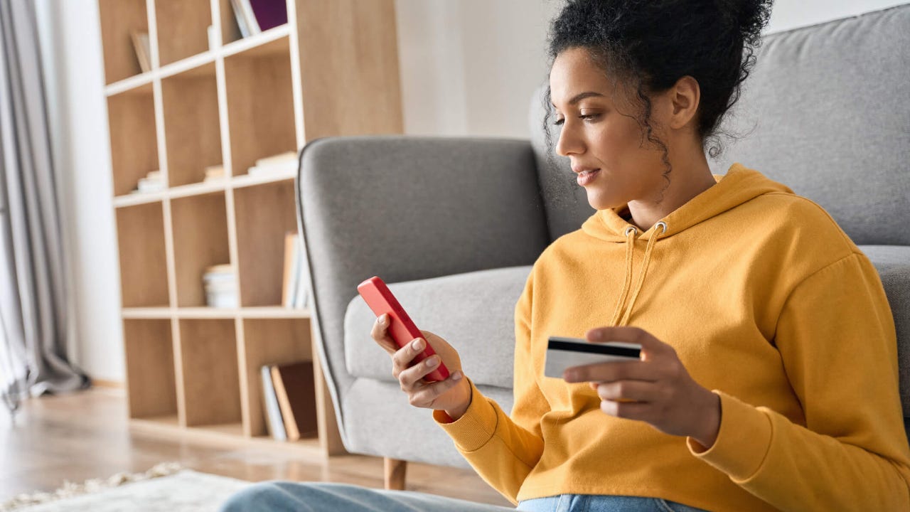 Young woman sits on the floor with her back against a couch as she uses her credit card to make a mobile purchase on her smartphone