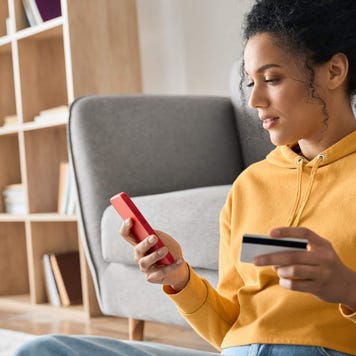 Young woman sits on the floor with her back against a couch as she uses her credit card to make a mobile purchase on her smartphone