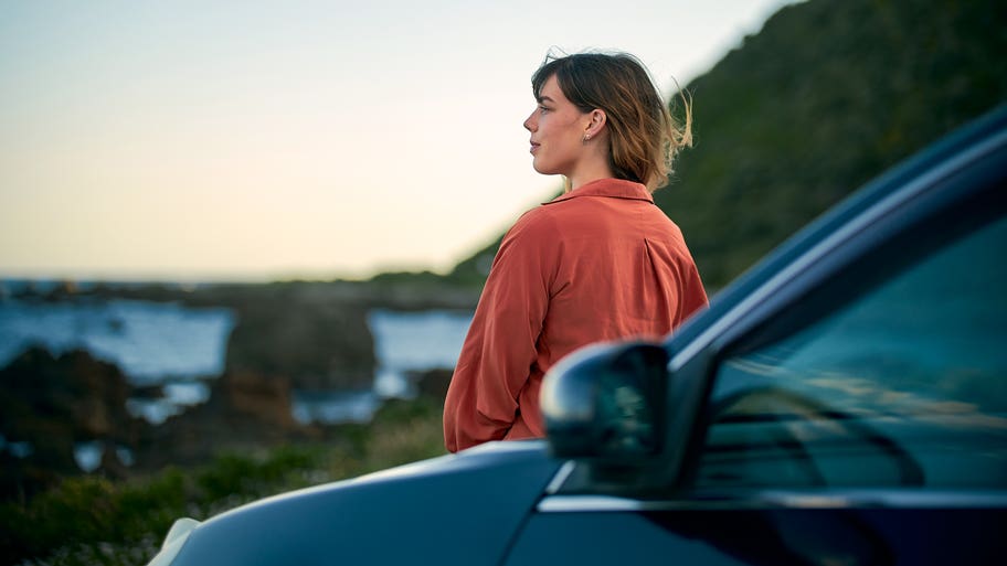 An adult stands by their car and stares out at a nice scene as the sun sets in the distance.