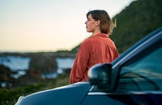 An adult stands by their car and stares out at a nice scene as the sun sets in the distance.