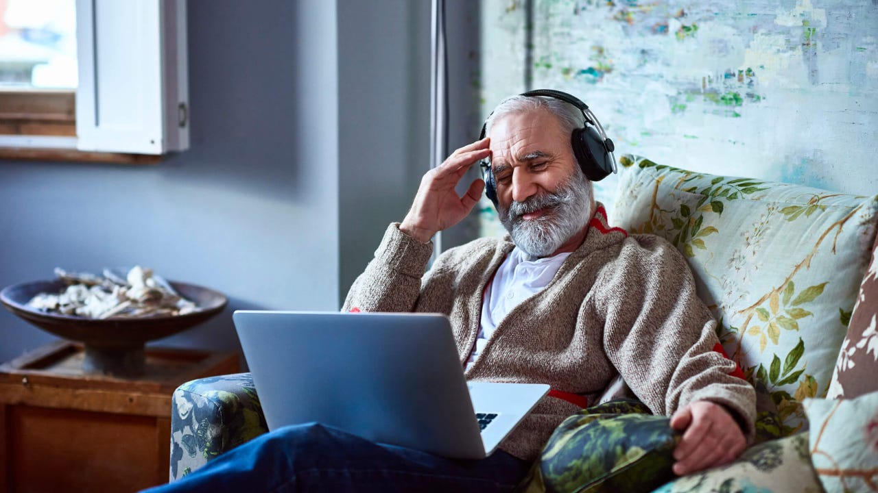 Senior man wearing headphones and using a laptop reclines in armchair as he streams a movie at home