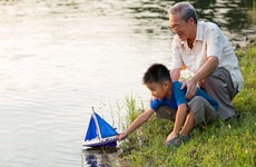 Grandparent and child are having a fun time together on the lakeshore where the child is putting a toy sailboat into the water.