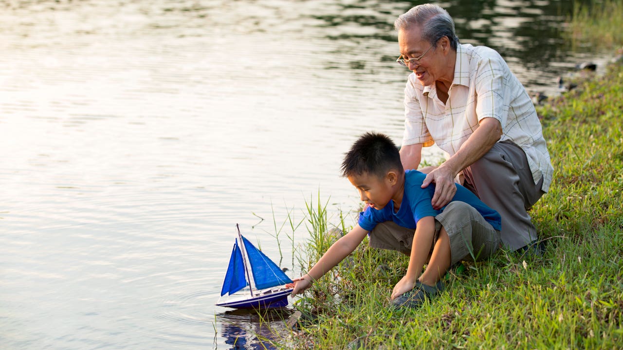 Grandparent and child are having a fun time together on the lakeshore where the child is putting a toy sailboat into the water.