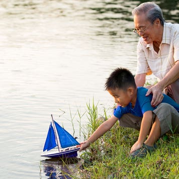 Grandparent and child are having a fun time together on the lakeshore where the child is putting a toy sailboat into the water.