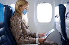 Young woman wearing mask in airplane seat looks at magazine