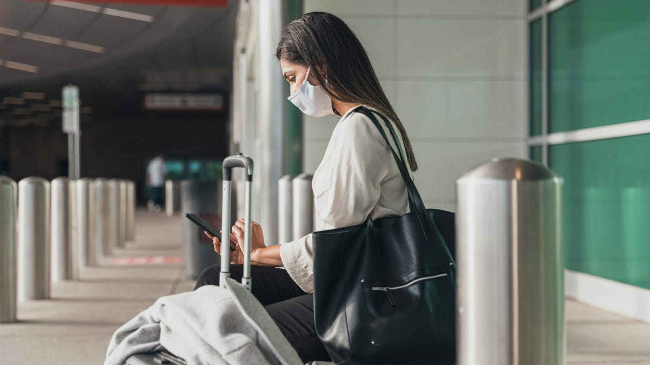Young woman sitting outside airport terminal and wearing a mask uses her smartphone while surrounded by her luggage