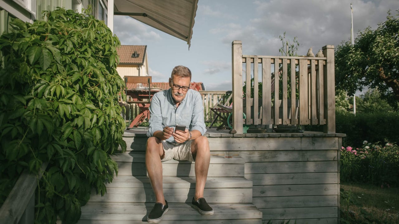 Senior man uses smartphone while sitting on his home's back porch step