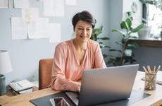 Middle aged woman smiles and uses laptop in home office at desk