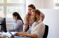 Two coworkers wearing masks in an office gather around a desktop computer and laptop