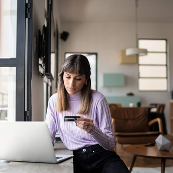 A young woman holds her credit card while working on a laptop in her apartment