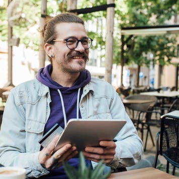 Young man at outdoor cafe smiles as he holds his tablet and credit card