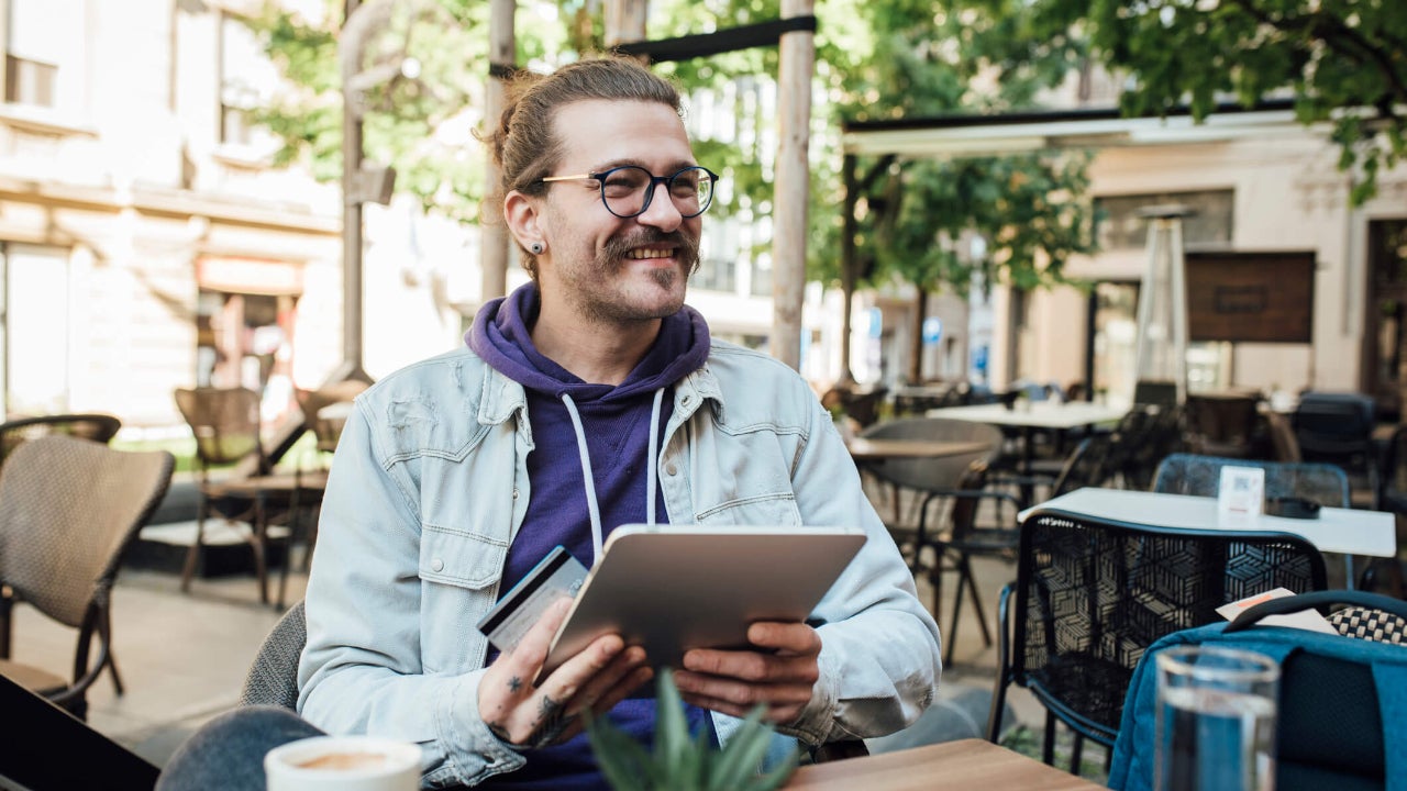 Young man at outdoor cafe smiles as he holds his tablet and credit card