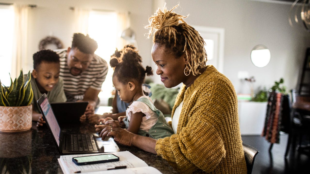 Black family, mother working from home holding toddler-age daughter, with husband and son in background