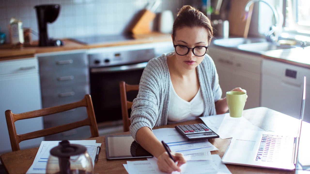 Young woman does finances at kitchen table with calculator, laptop, and sheets of paper spread out