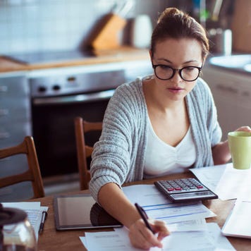 Young woman does finances at kitchen table with calculator, laptop, and sheets of paper spread out