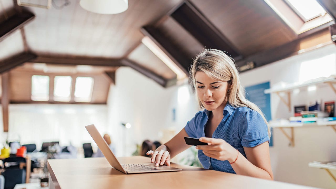 Young woman at a shared workspace uses her laptop while looking down at her credit card