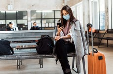 woman witha. mask sitting at an airport terminal with her phone in hand