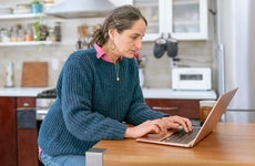 A mid adult woman sits at the counter in her kitchen and uses a laptop computer for online banking while managing her personal finances.