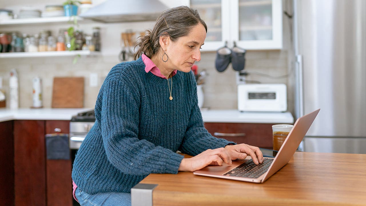 A mid adult woman sits at the counter in her kitchen and uses a laptop computer for online banking while managing her personal finances.