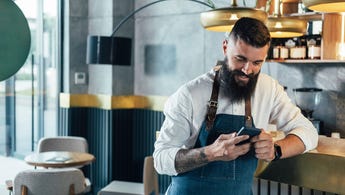 Young adult male wearing an apron at a cafe smiles while using his smartphone