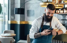 Young adult male wearing an apron at a cafe smiles while using his smartphone